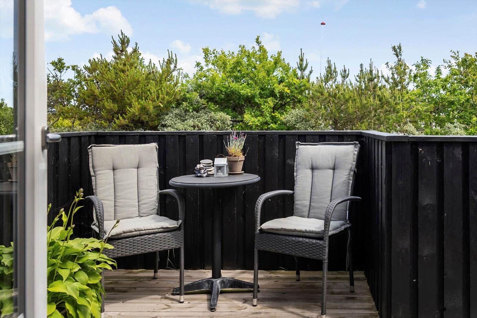Terrasse mit zwei Stühlen und Tisch, umgeben von grüner Vegetation und dunklem Holzzaun.