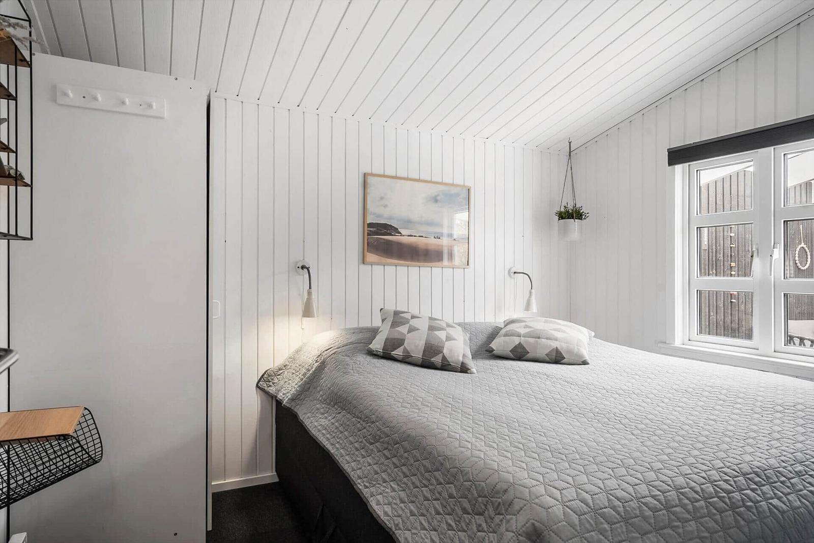 Schlafzimmer mit weiß verkleideten Wänden, Bett mit grauer Decke und Fenster.