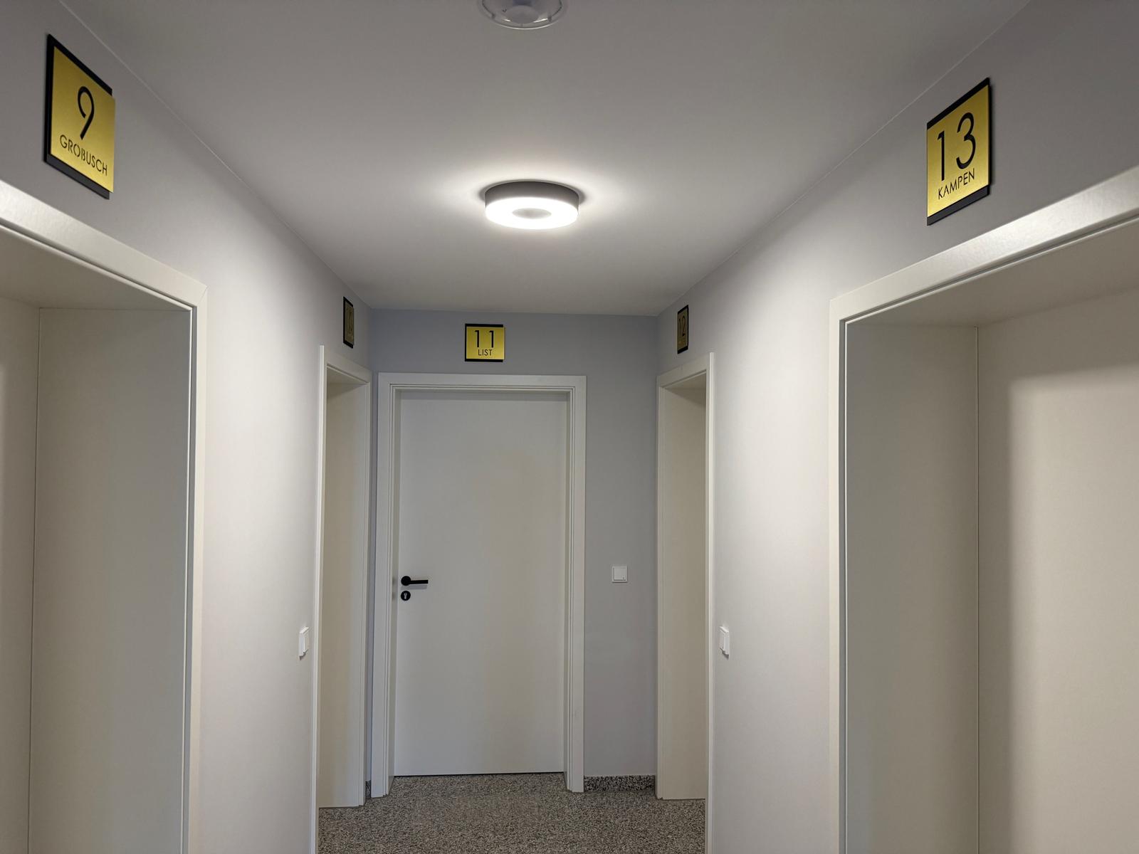 Flur mit Türen und gelben Schildern mit Zimmernummern und Namen.