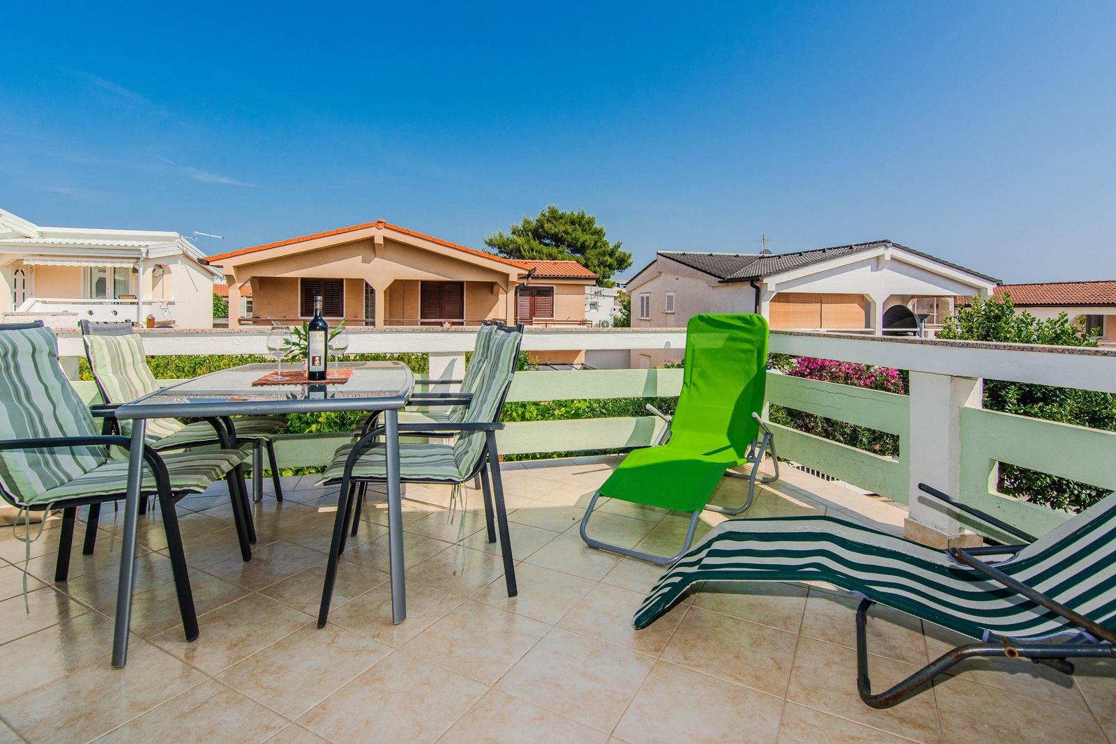 Terrace with table, chairs, and loungers. Background: houses and flowers.