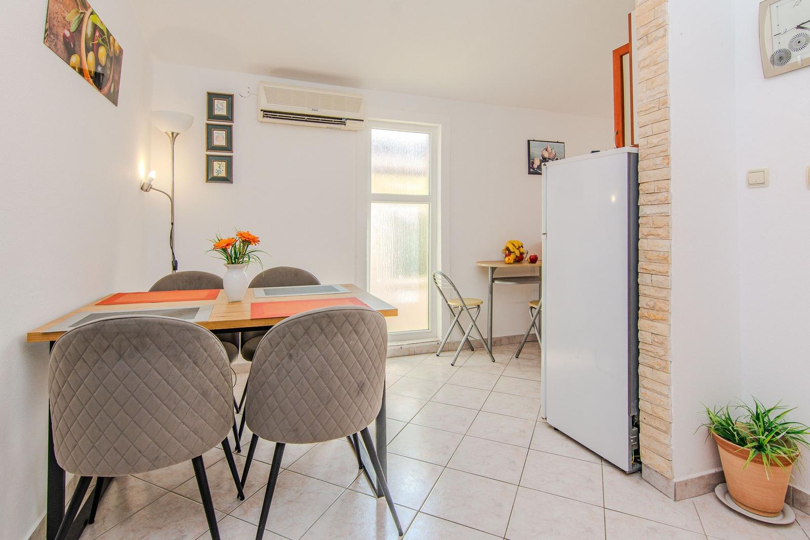 Dining area with table, chairs, and refrigerator. Window and plants visible.