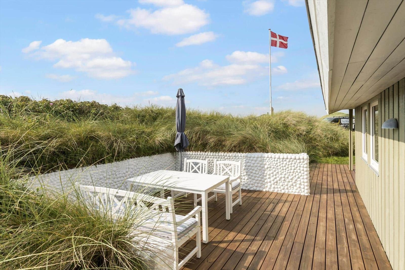 Terrasse mit weißem Tisch und Stühlen, daneben Flagge und Strandvegetation.