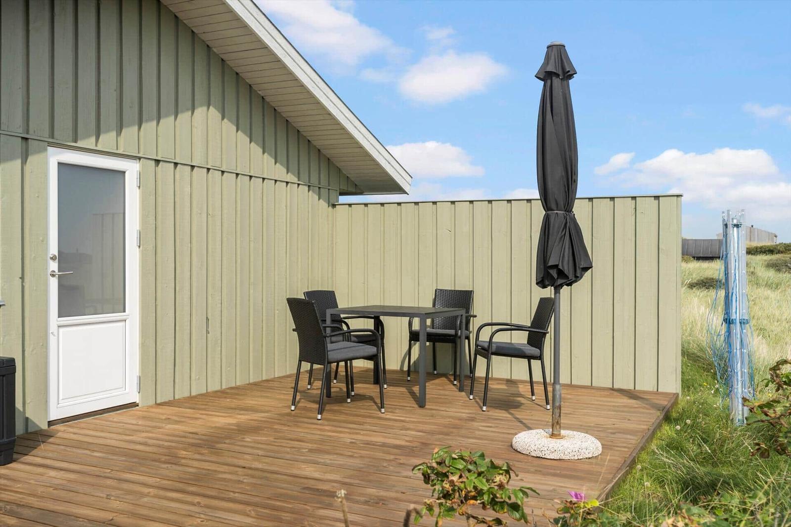 Terrasse mit Tisch, vier Stühlen und geschlossenem Sonnenschirm. Holzdeck und grüne Umgebung.