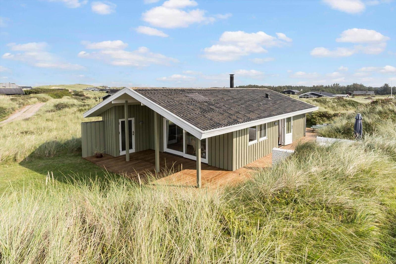 Moderne Hütte mit Terrasse in Dünenlandschaft unter blauem Himmel.
