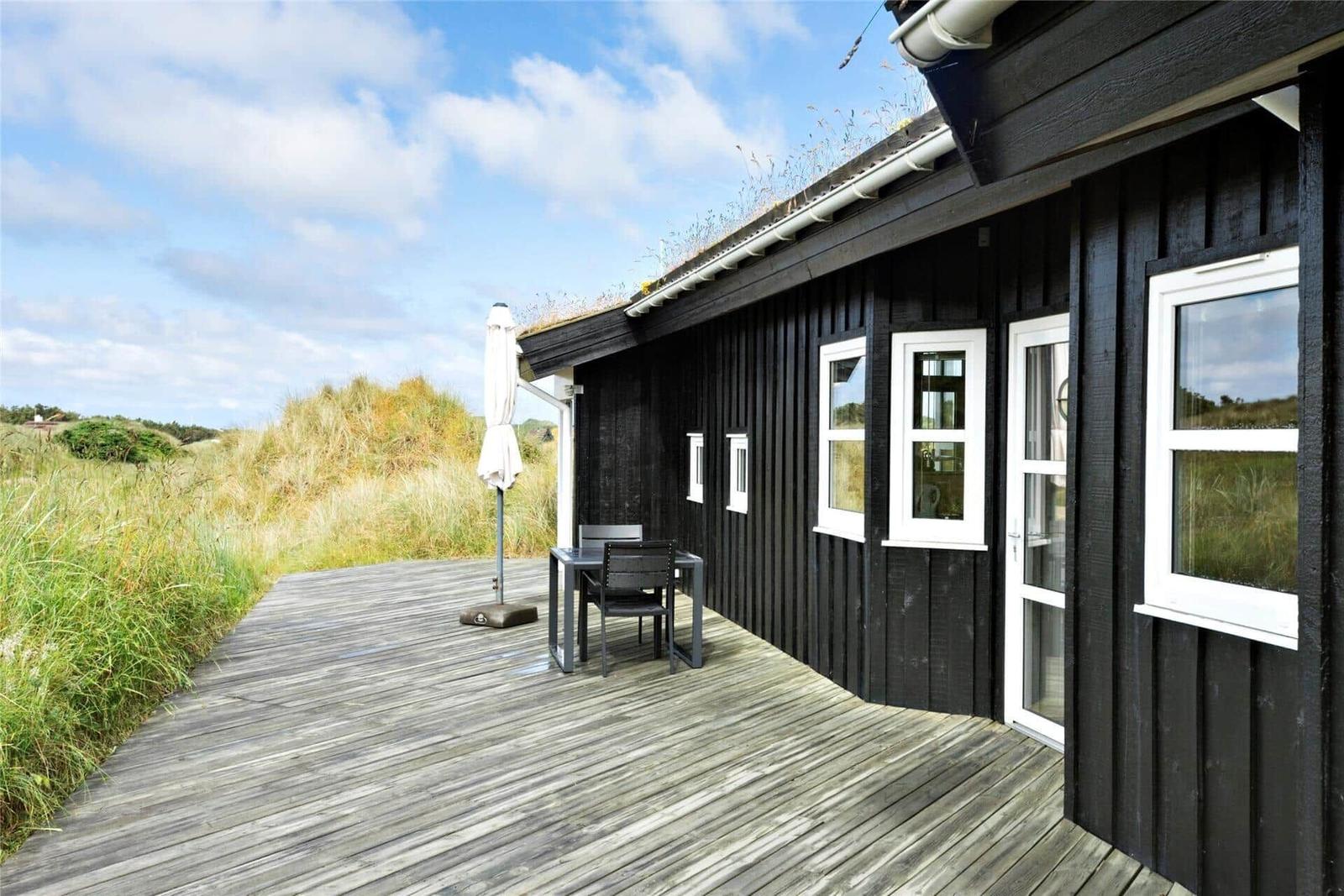 Schwarzes Holzhaus mit Terrasse, Tisch, Stühlen und Sonnenschirm. Grasland im Hintergrund.