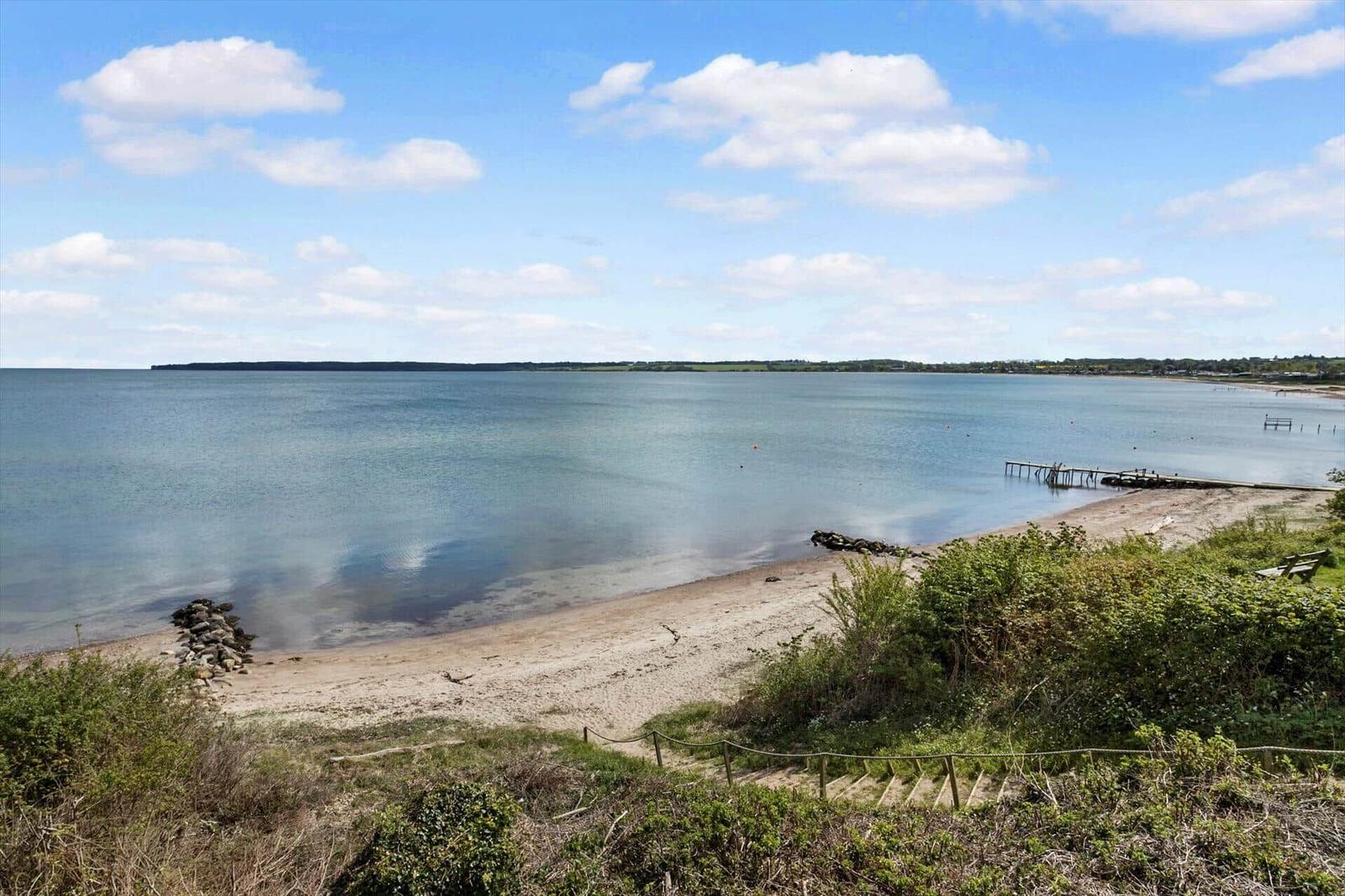 Strandufer mit Steg und sanfter Bucht unter blauem Himmel.