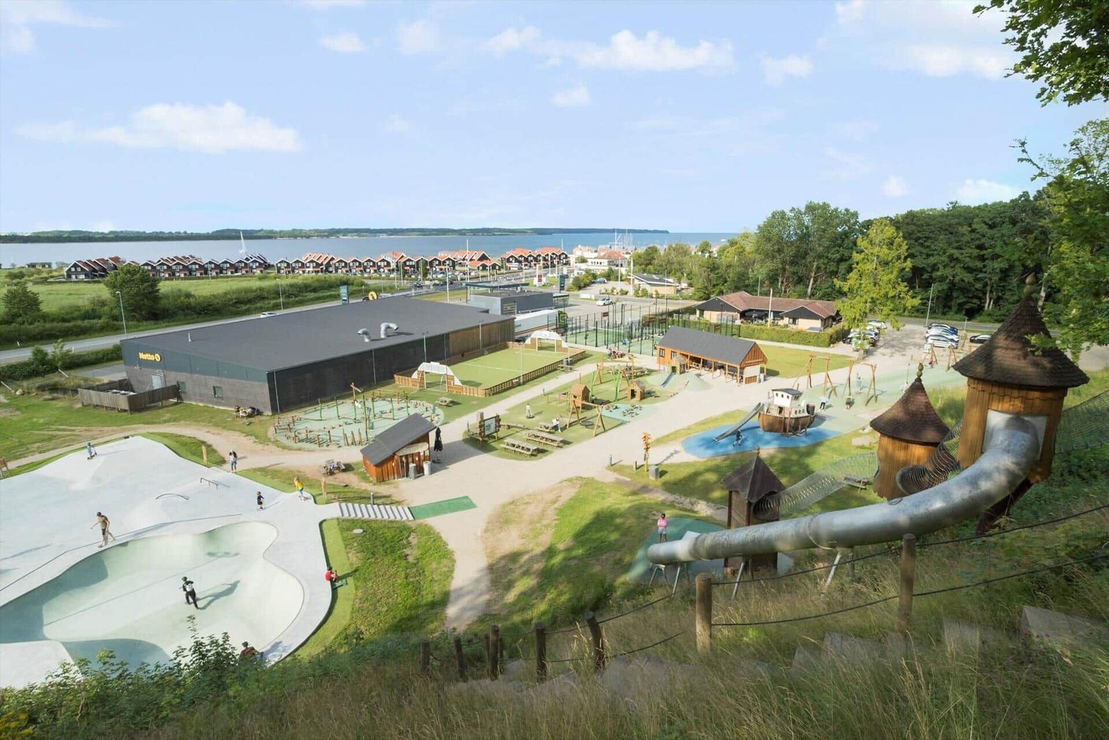 Großer Spielplatz mit Rutsche, Klettergerüst und Skatepark neben Wohngebäuden am Wasser.