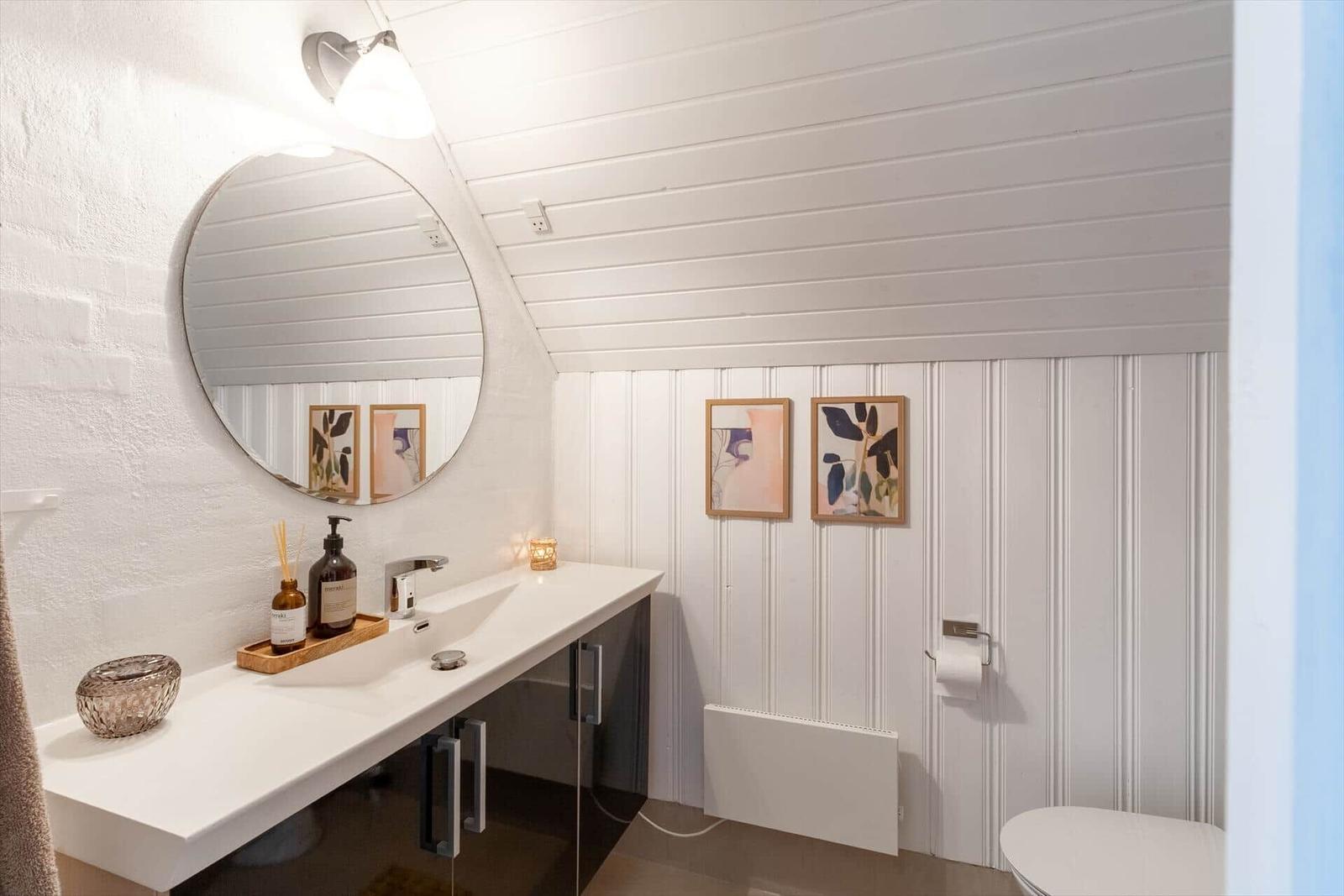 Badezimmer mit Waschbecken, Spiegel und Toilette. Weiße Wände und Holzvertäfelung.