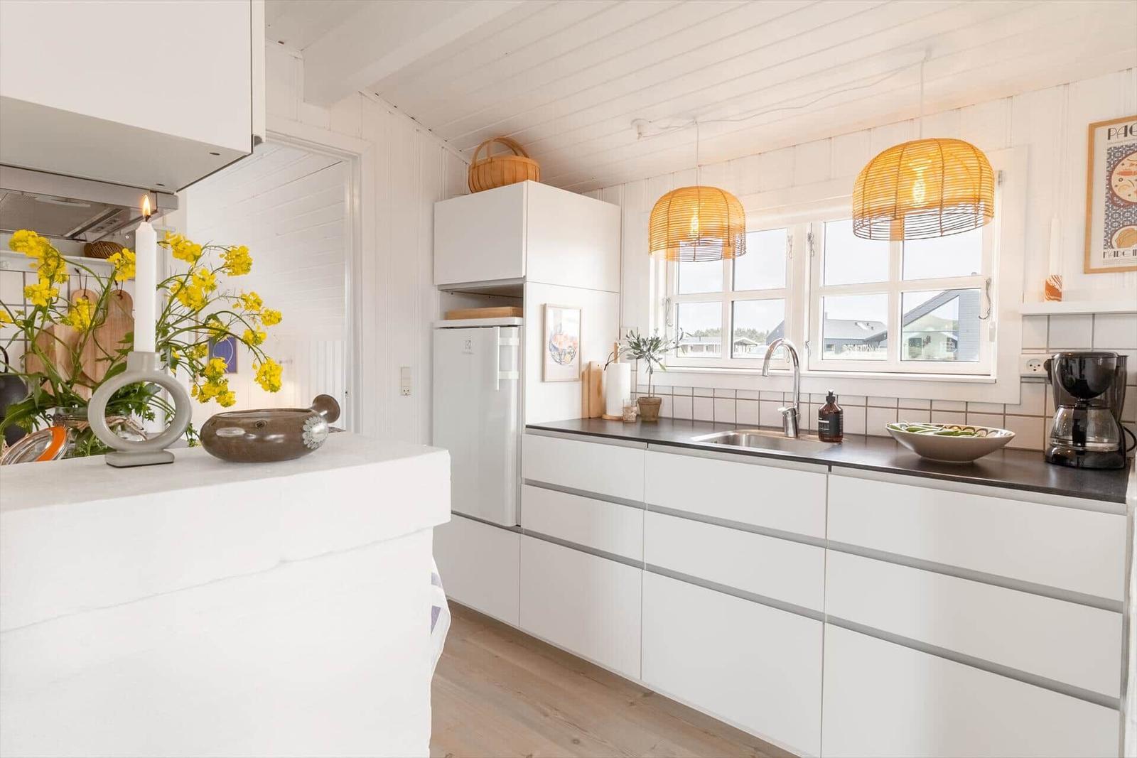 Küche mit weißen Schränken, Arbeitsplatte und Fenster. Hängelampen und Blumen sind sichtbar.