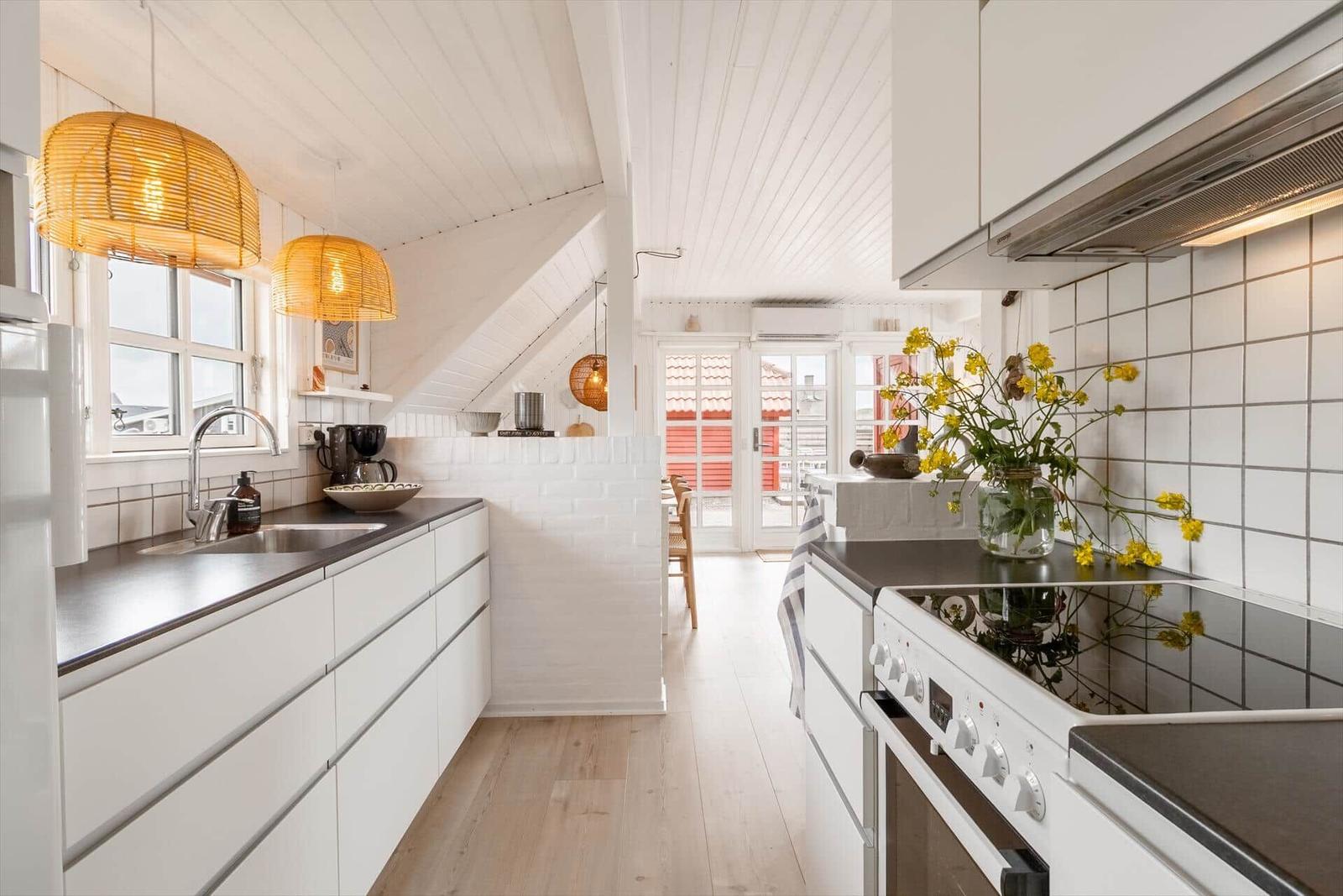 Küche mit weißen Schränken, dunklen Arbeitsflächen und gelben Lampen. Blick in Wohnbereich mit Holzboden.