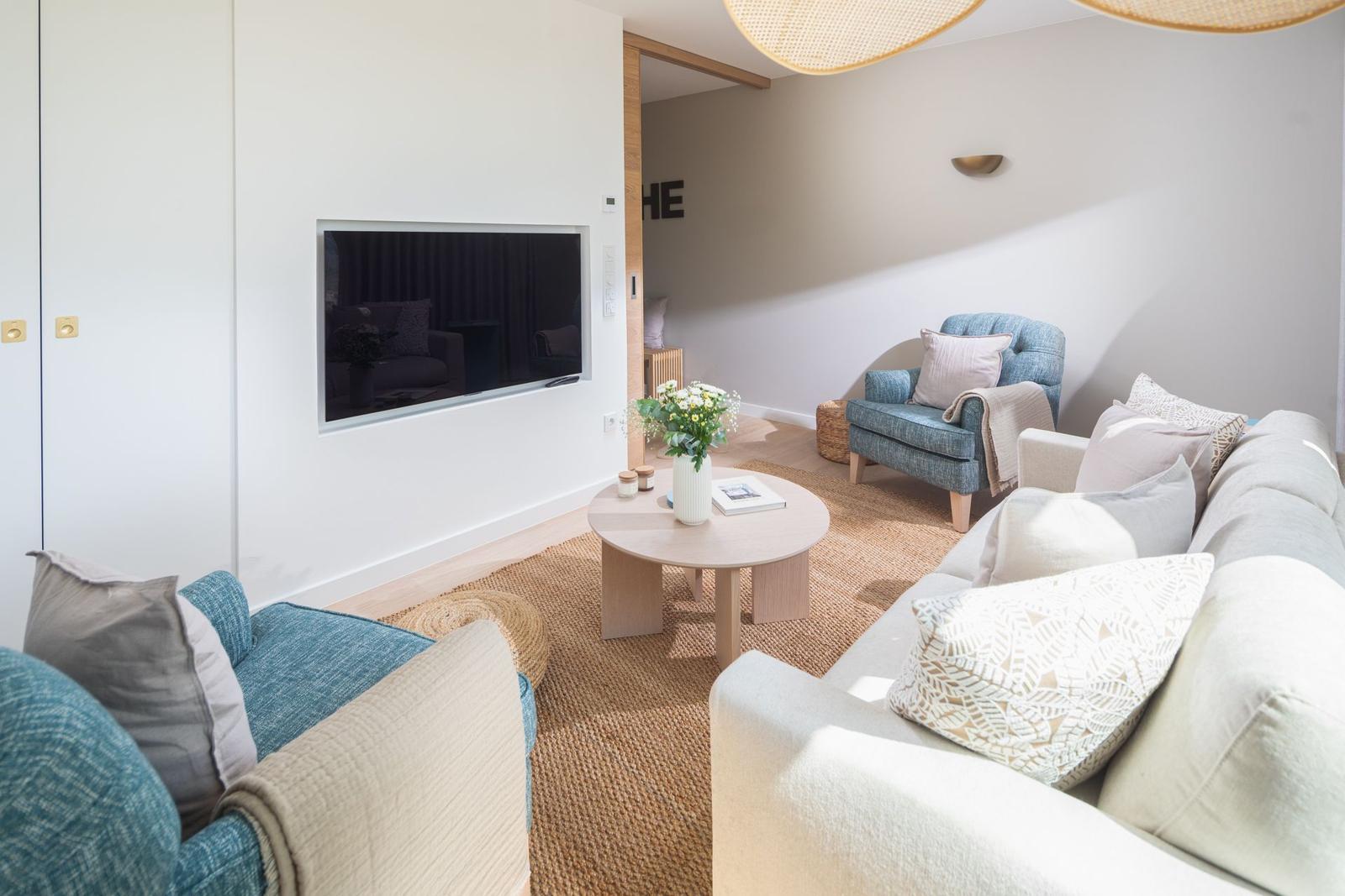 Wohnzimmer mit Sofa, Sessel und runden Tisch. Fernseher an Wand. Teppich und Beleuchtung.