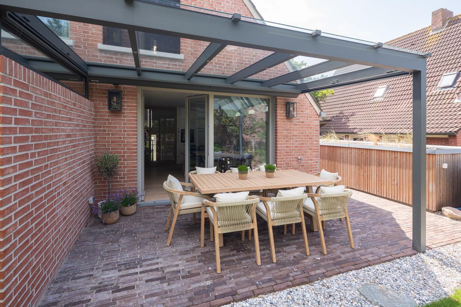 Terrasse mit Holztisch, Stühlen und Glasdach. Roter Backsteinbau und Pflanzen sichtbar.