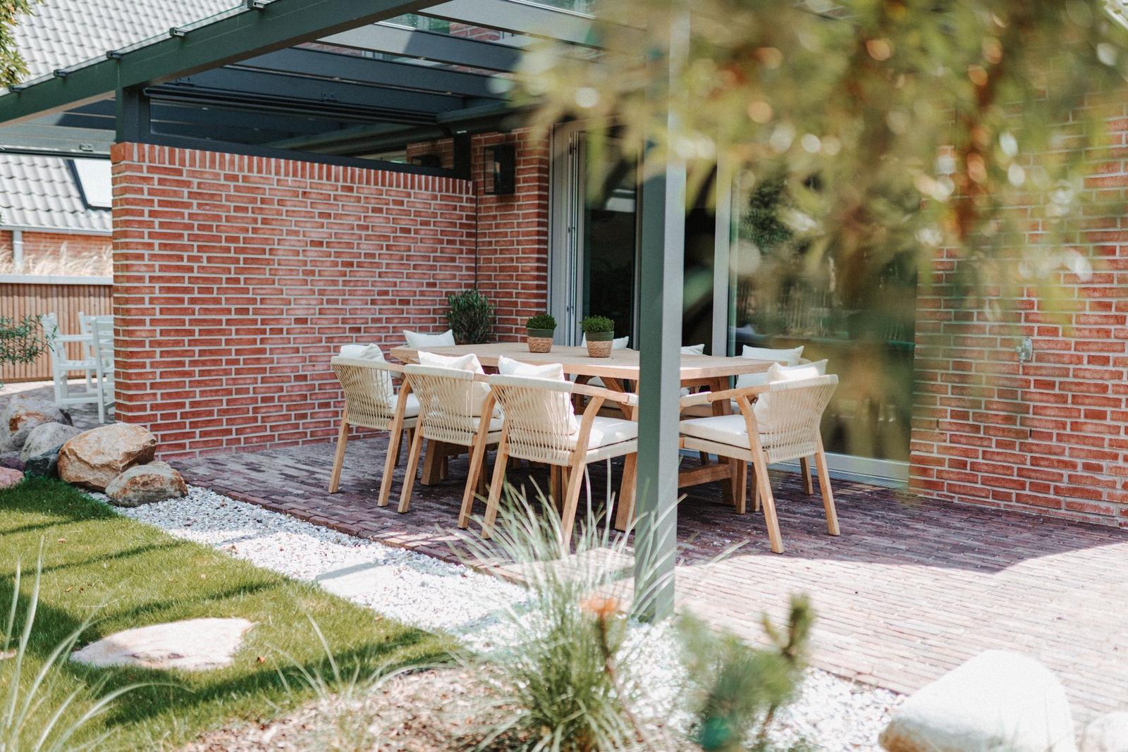 Terrasse mit Holztisch, Stühlen und Pflanzen unter Glasdach.