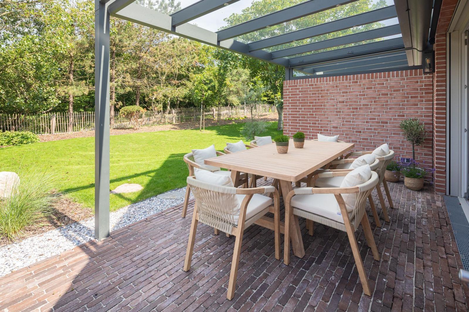 Terrasse mit Tisch und Stühlen unter Pavillon. Hintergrund: grüner Garten und Backsteinmauer.