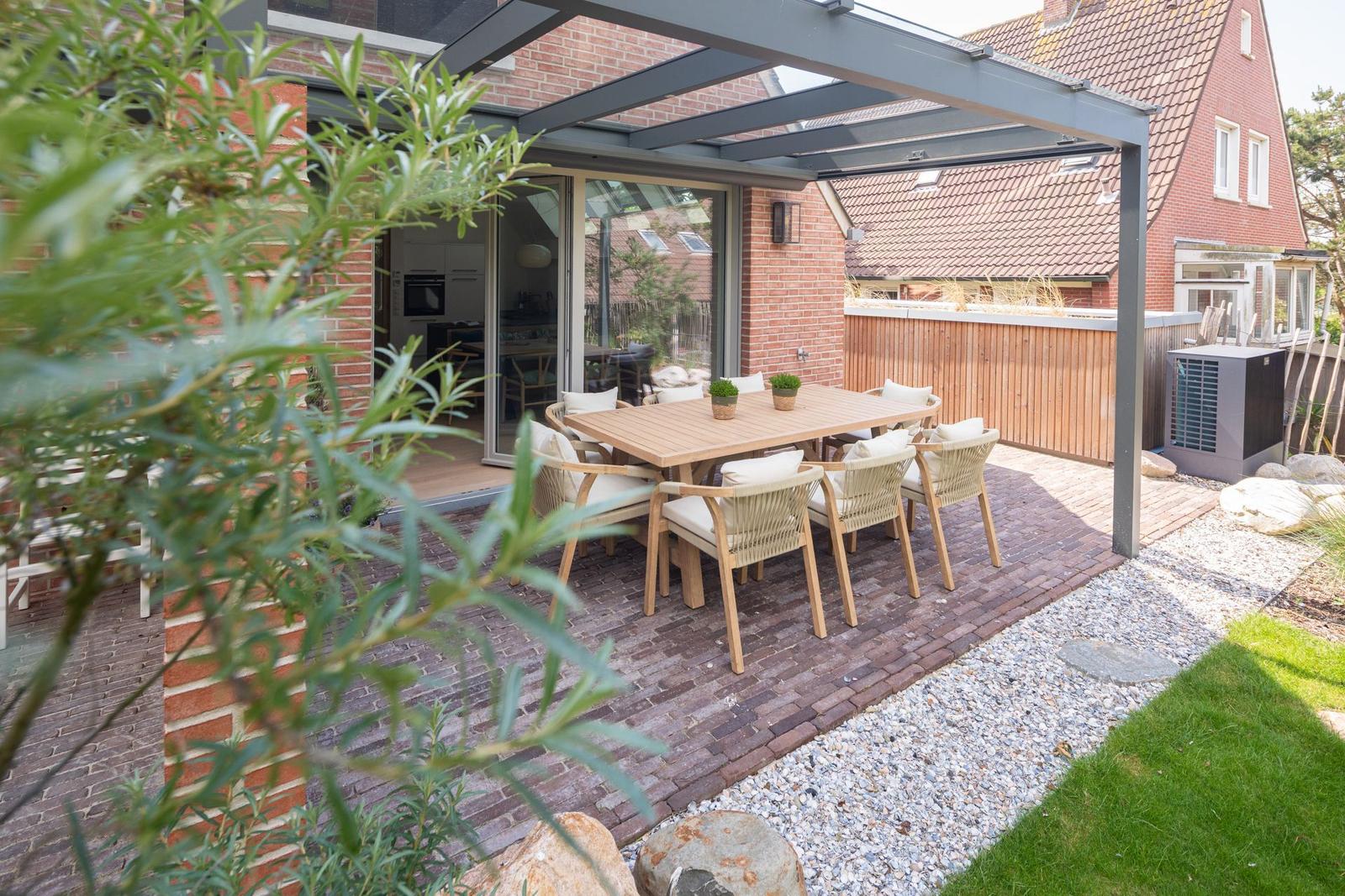 Terrasse mit Holztisch und Stühlen unter Glasdach. Hintergrund: Haus mit rotem Ziegeldach.