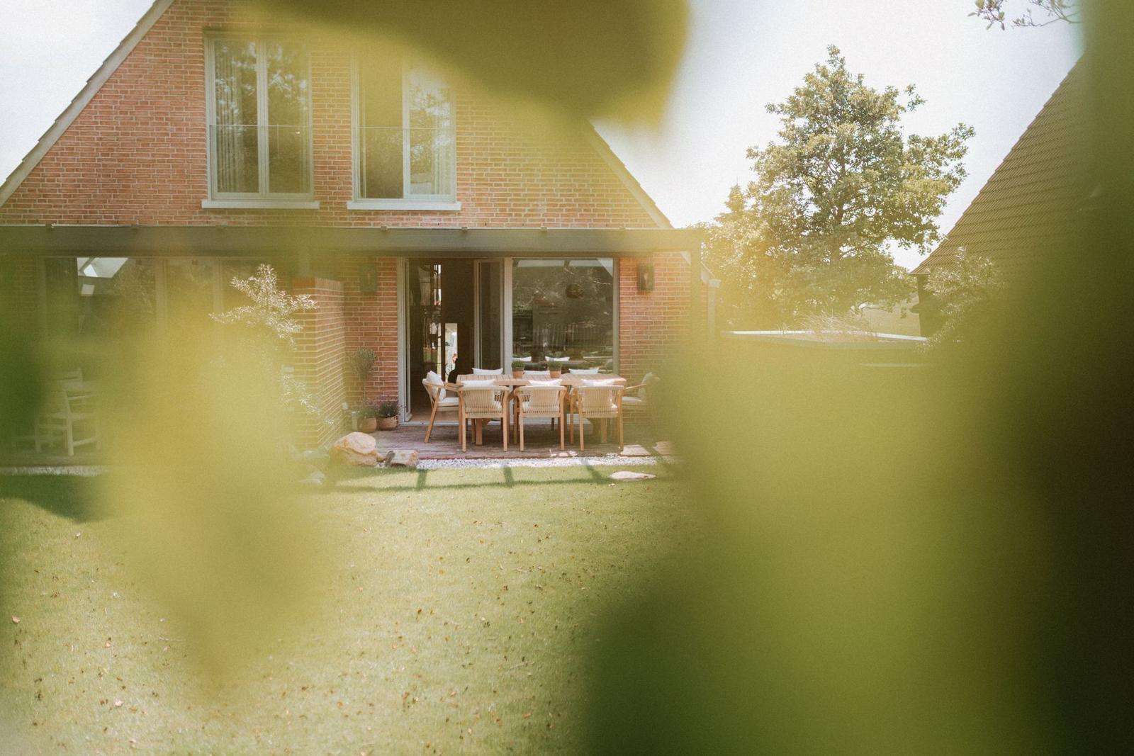 Haus mit Terrasse und Garten. Holztisch mit Stühlen. Blick durch Blätter.