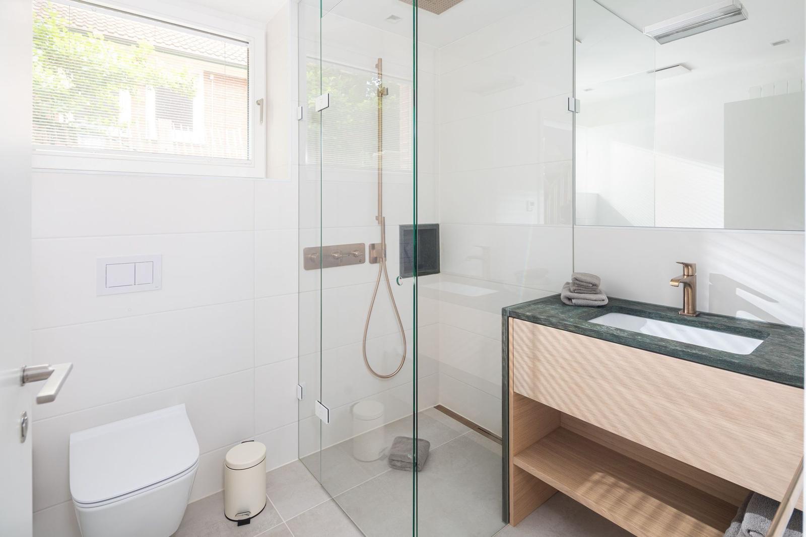 Bad mit Dusche, Waschbecken und Toilette. Glaswand, Holzunterschrank und Fenster.