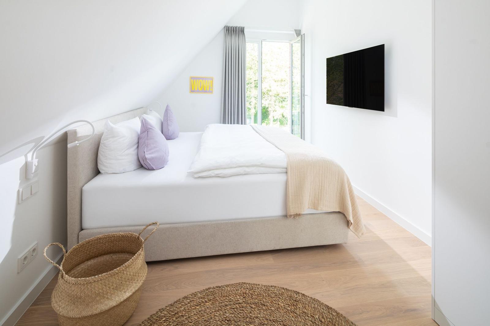 Schlafzimmer mit Bett, TV und Fenster. Holzboden und Wandlampe sichtbar.