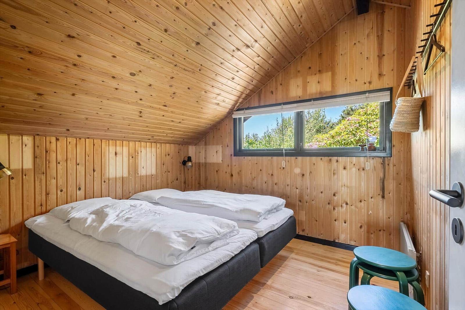 Schlafzimmer mit Doppelbett, Holzwand und Fensterblick auf Grün.