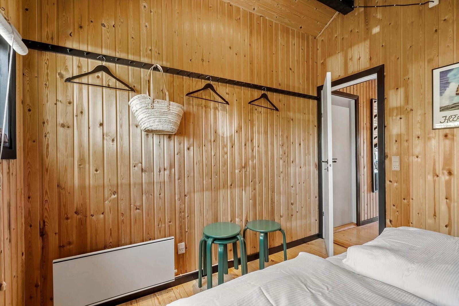 Zimmer mit Holzwand, Bett, zwei grünen Hockern und Kleiderhaken.