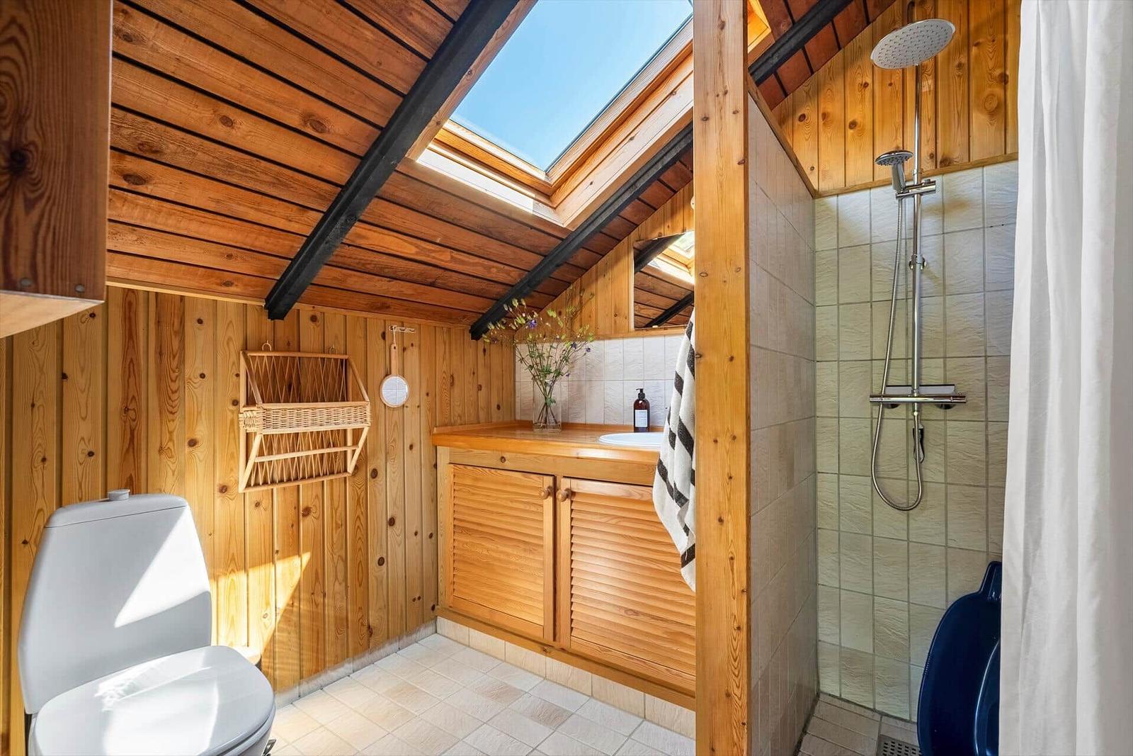 Badezimmer mit Dusche, Waschbecken und Toilette unter Holzdecke mit Dachfenster.
