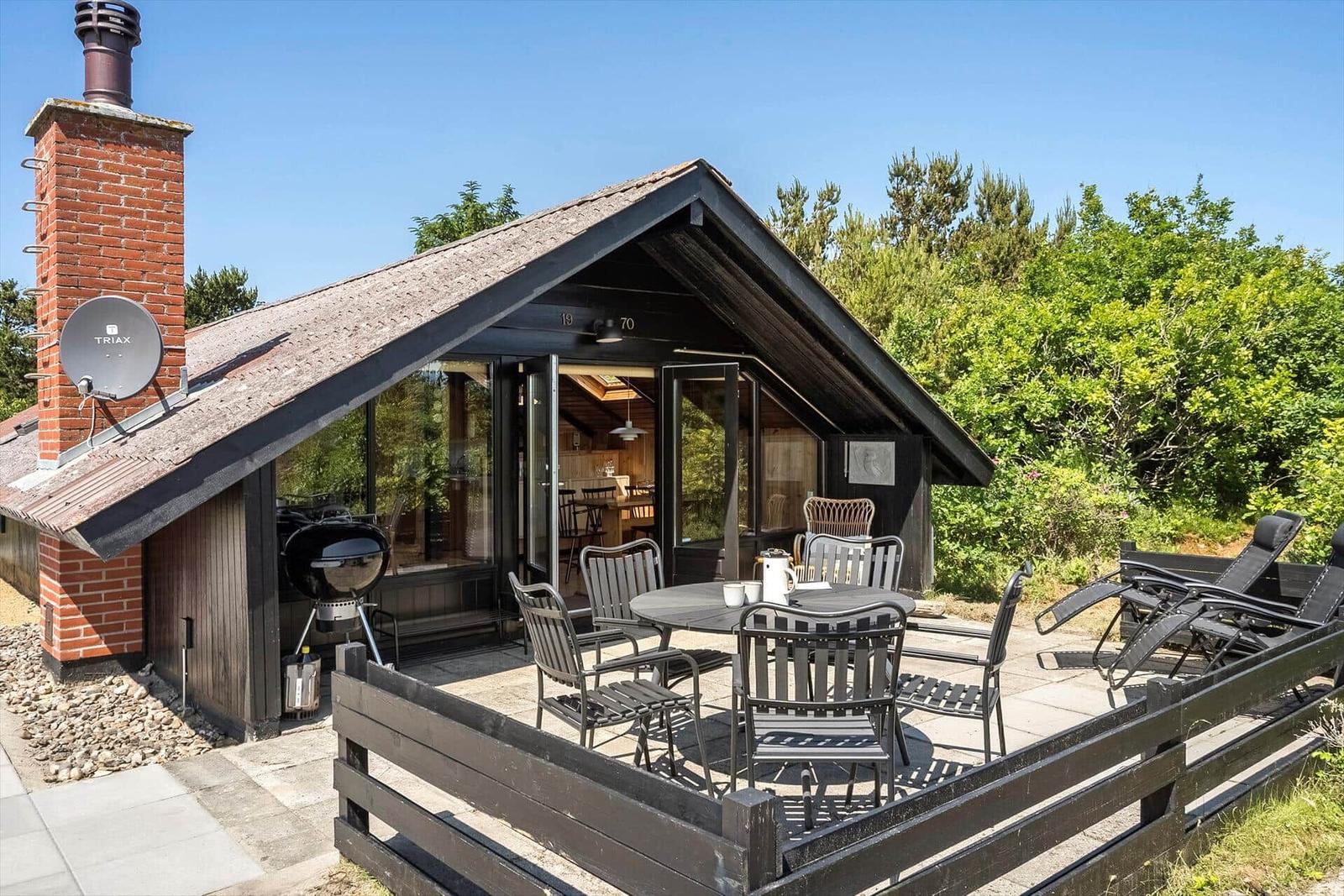 Haus mit Terrasse, Grill und Sitzgelegenheiten. Holzhaus mit Schiebetüren und Satellitenschüssel.