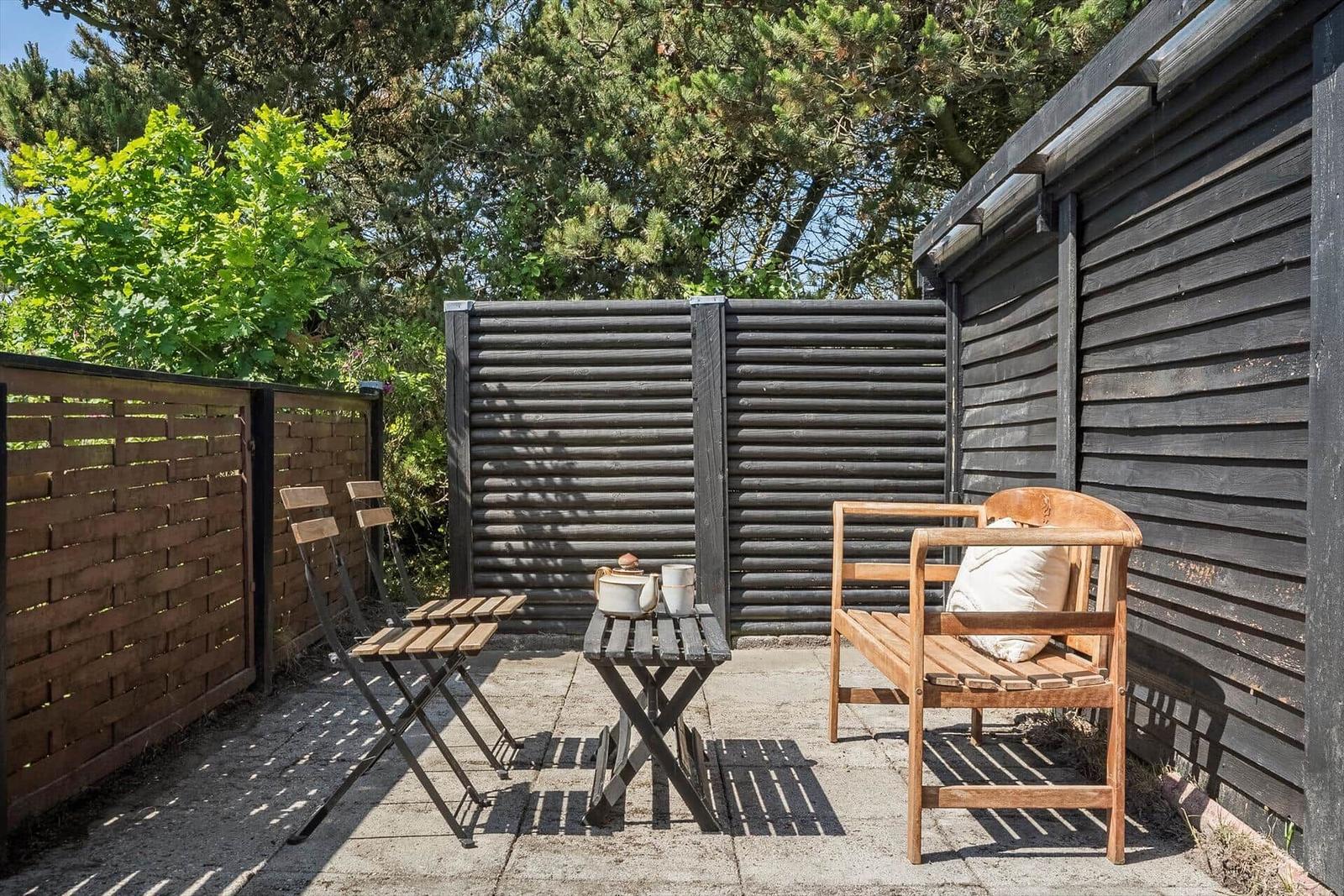 Terrasse mit Holzbank, Tisch und Stühlen. Umgeben von Holzwänden und Bäumen.