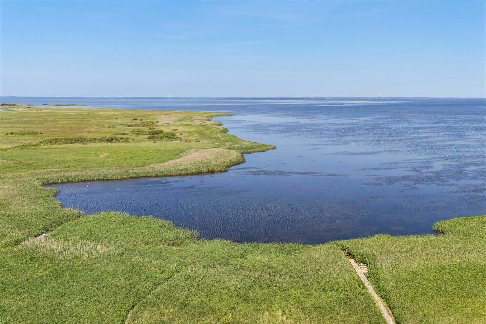 Weitläufiger Blick auf grüne Wiesen und ruhiges Wasser unter blauem Himmel.