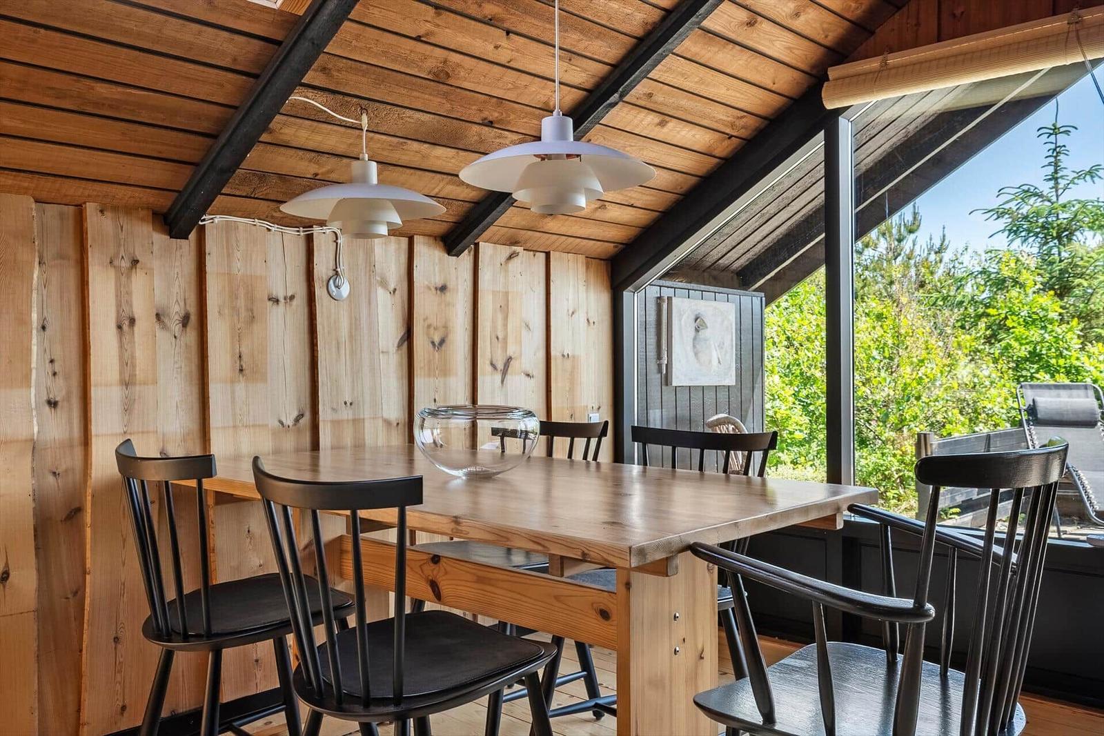 Essbereich mit Holztisch, schwarzen Stühlen und Holzwand. Große Fenster mit Blick auf Grün.