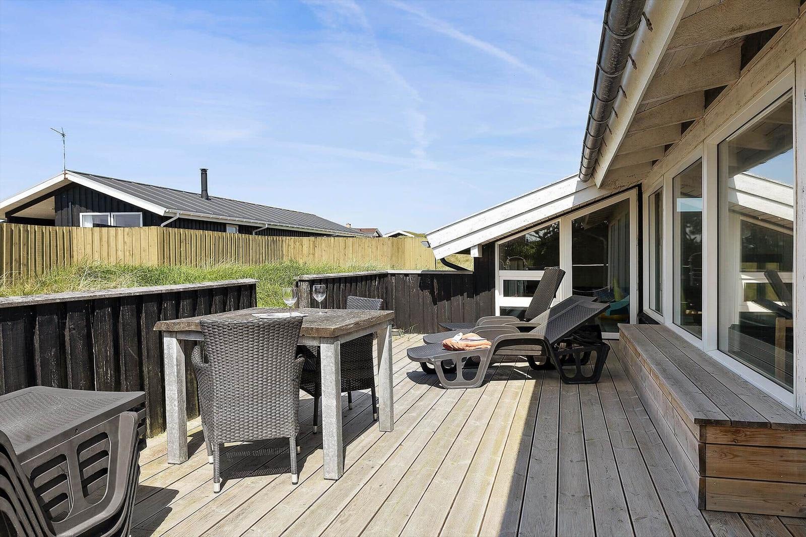 Terrasse mit Tisch, Stühlen und Liegen. Holzdecke, Fenster und Zaun sichtbar.