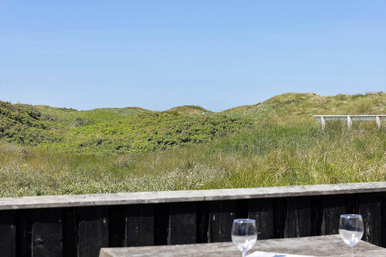 Terrasse mit Tisch und Gläsern, Blick auf grüne Dünen und klaren Himmel.