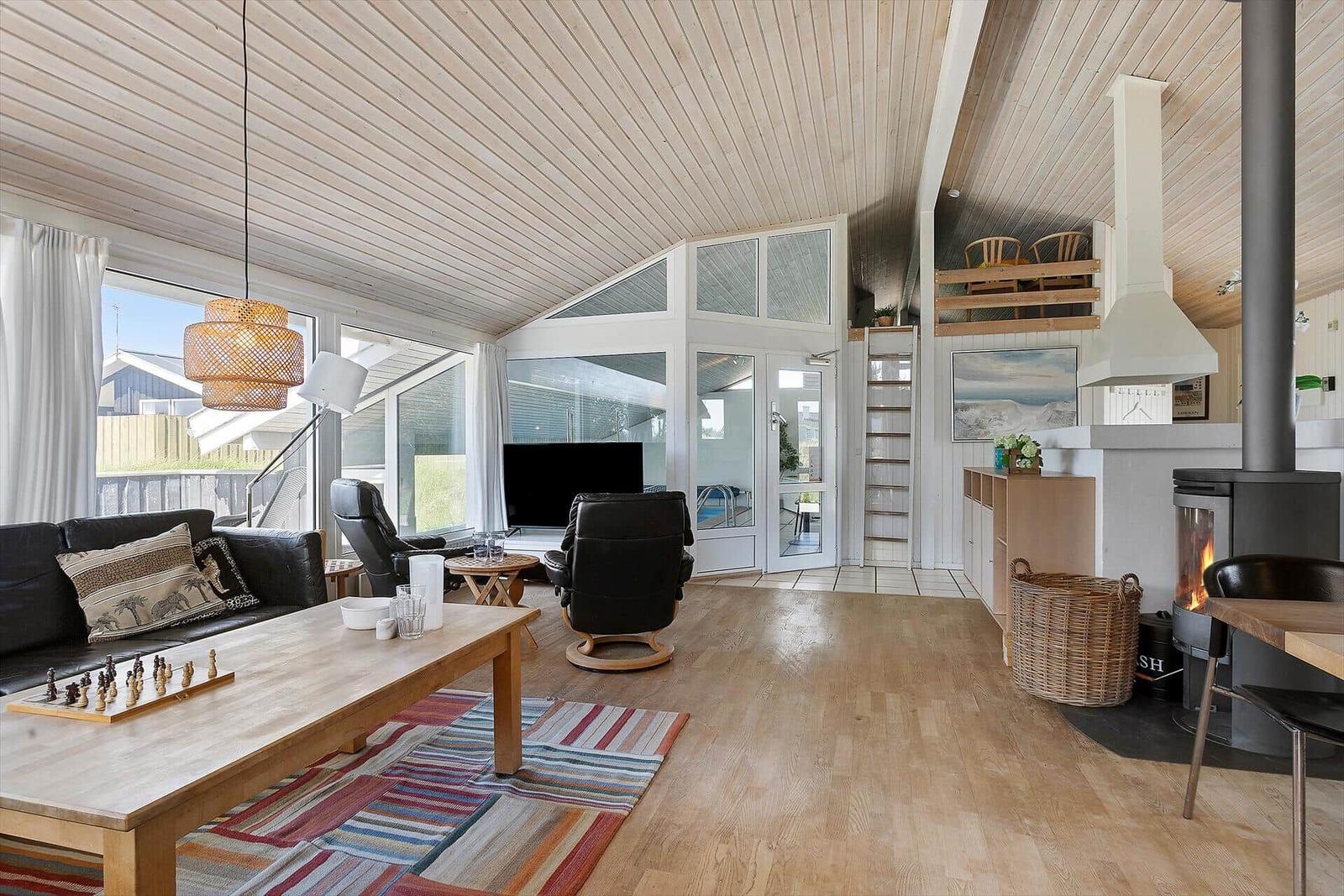 Wohnraum mit Holzboden, Sofa, Sessel und Kaminofen. Große Fensterfront mit Blick nach draußen.