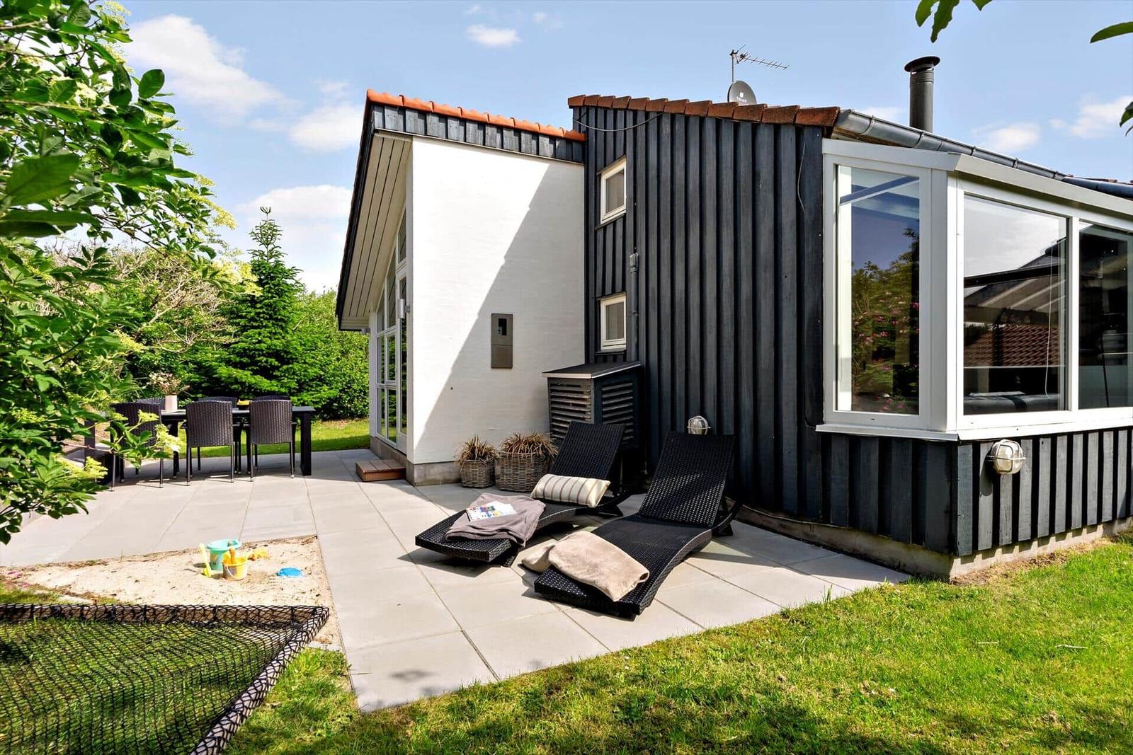 Terrasse mit Liegestühlen, Tisch und Garten. Schwarze Holzwand und großes Fenster.
