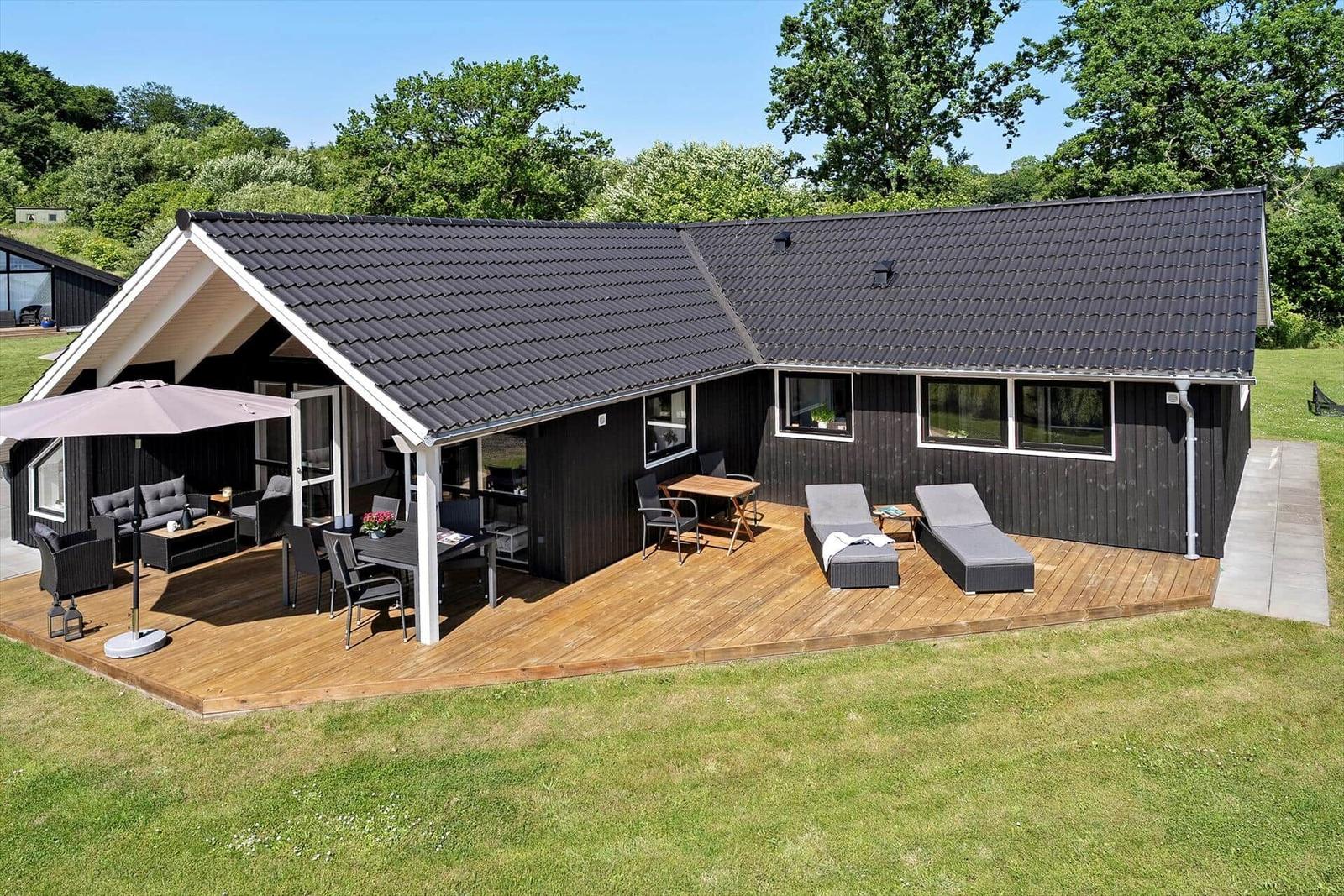 House with large wooden terrace, garden furniture, and view of green trees.