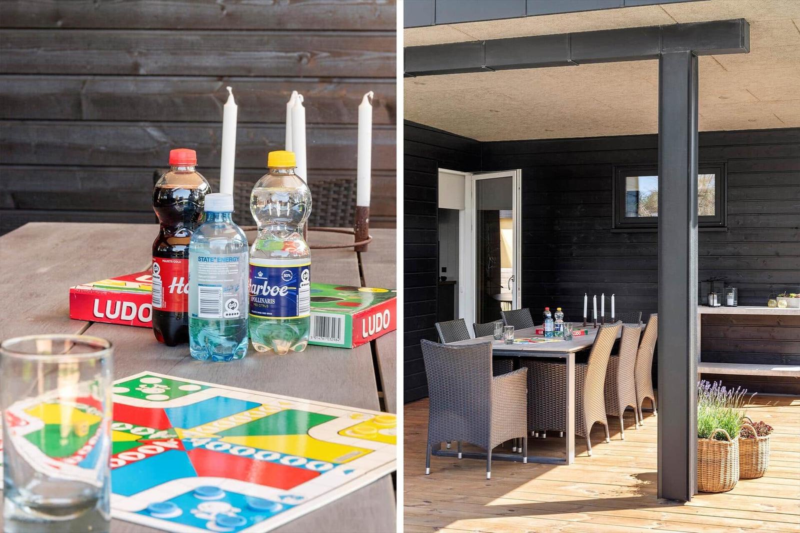 Terrasse mit Tisch, Stühlen und Ludo-Spiel. Hintergrund: schwarze Holzwand und Fenster.