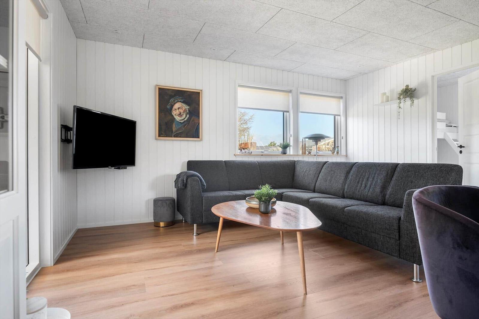 Wohnzimmer mit grauem Sofa, Holztisch und Wandfernseher. Fenster mit Blick nach draußen.