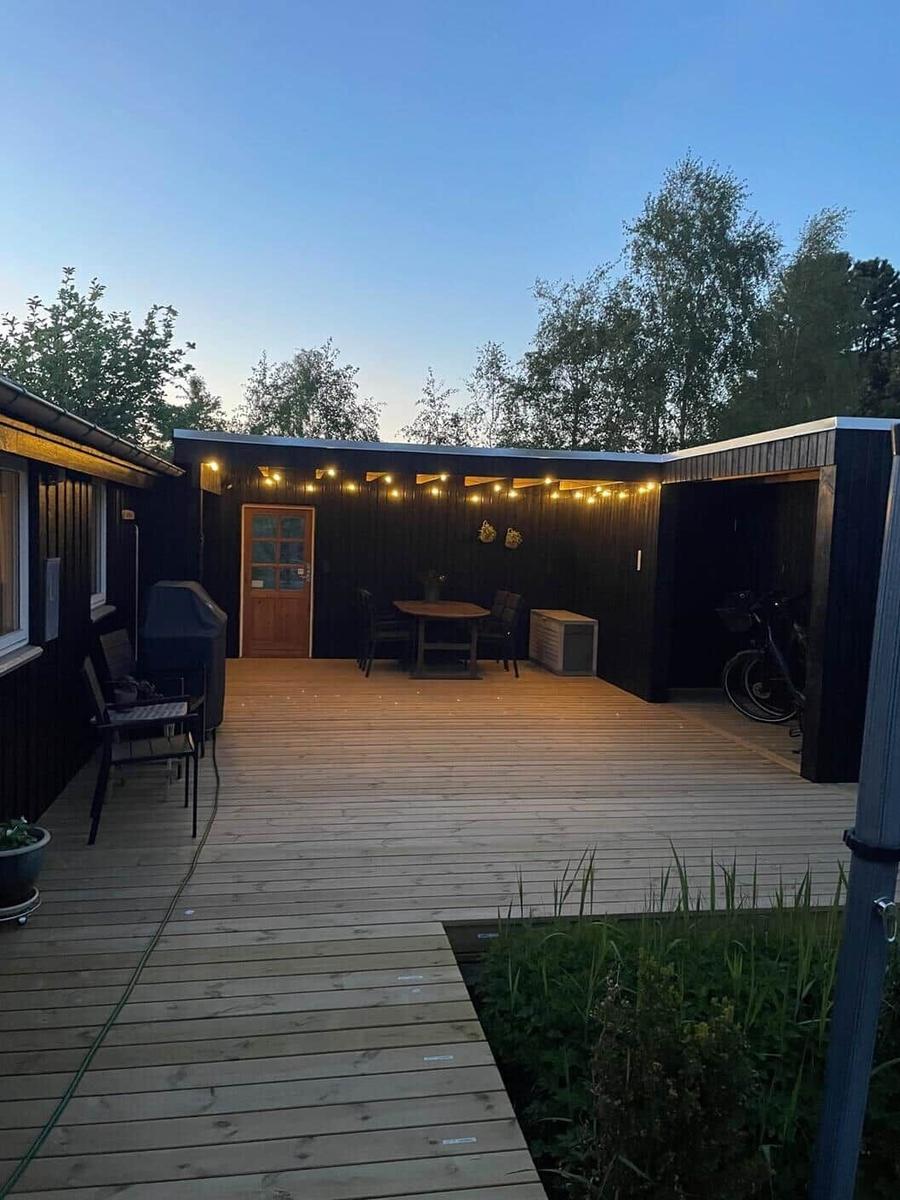 Holzterrasse mit Tisch, Stühlen und Grill. Hintergrund: Wald und Lichterkette.