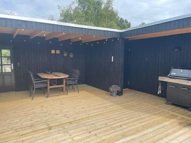 Terrasse mit Holzdecke, Tisch, Stühlen und Grill. Hintergrund: dunkle Holzwände und Bäume.