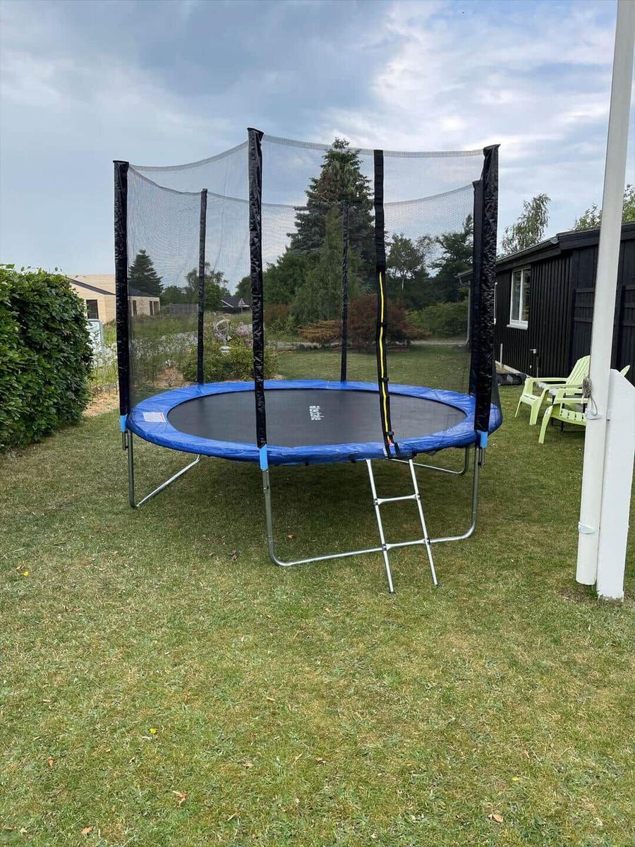 Ein Trampolin mit Sicherheitsnetz steht im Garten. Neben ihm befinden sich grüne Stühle.