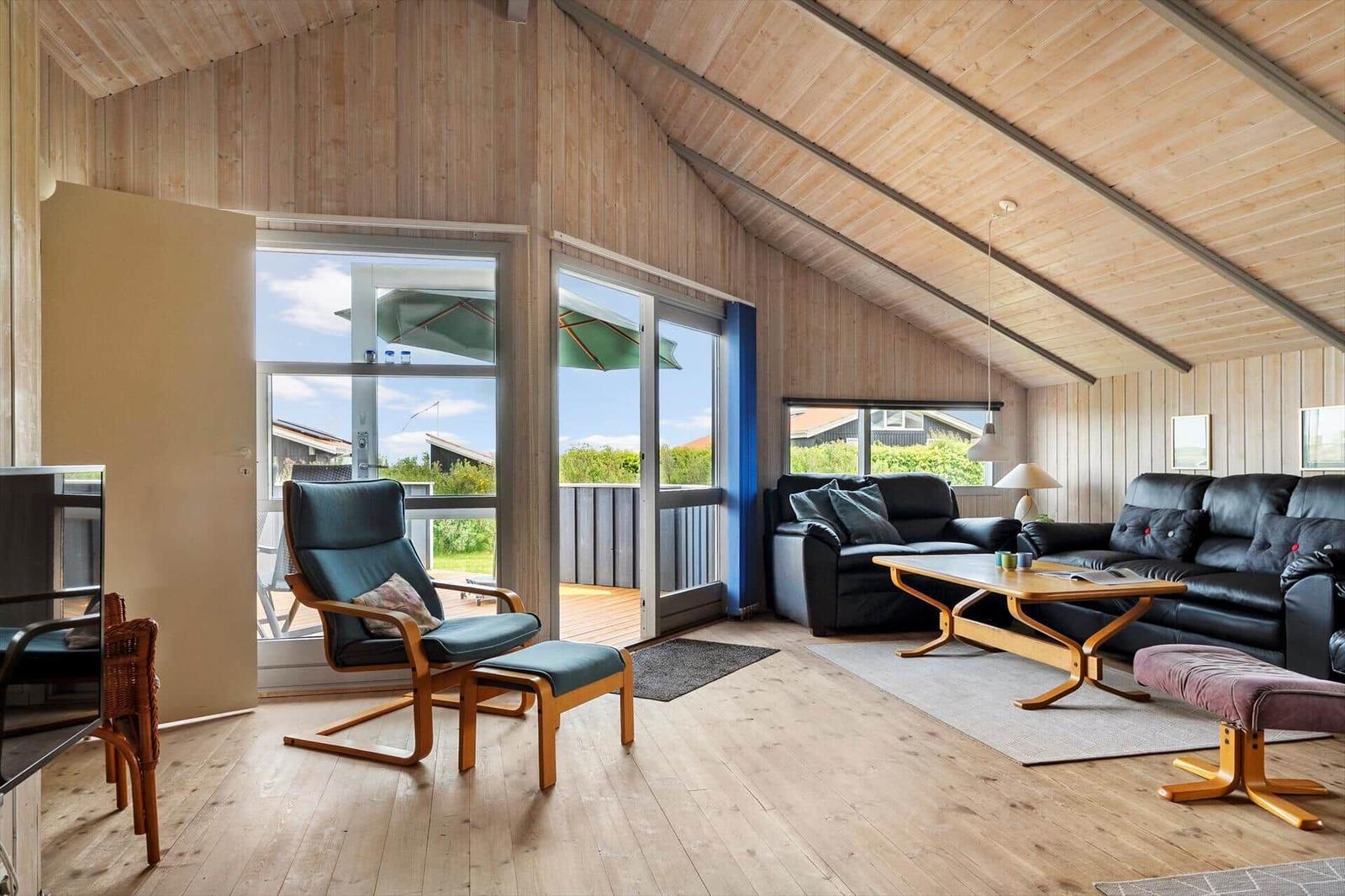 Living room with wooden ceiling, sofa and chair. Terrace with view of garden.