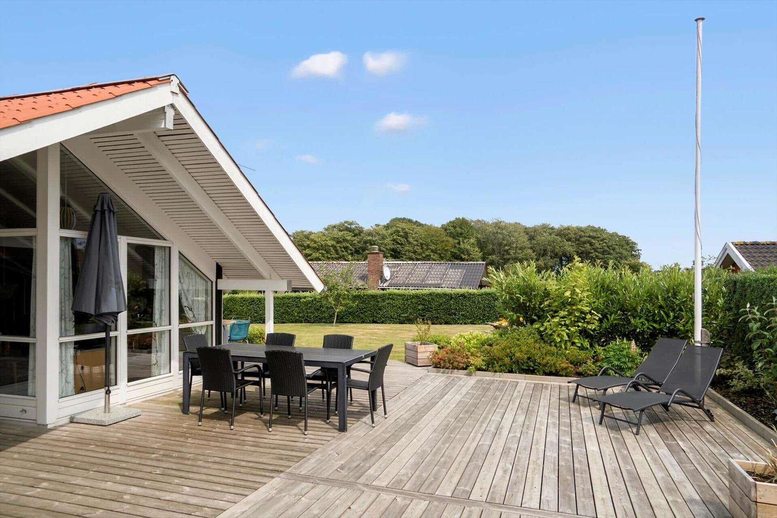 Deck with table, chairs, and lounge chairs. Backyard with green areas and hedges.