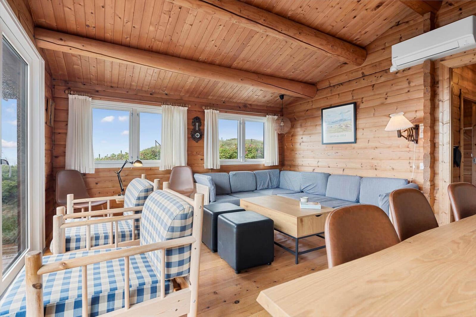 Wohnzimmer mit Holzwand, Sofa, Tisch und Stühlen. Fenster mit Blick auf Landschaft.