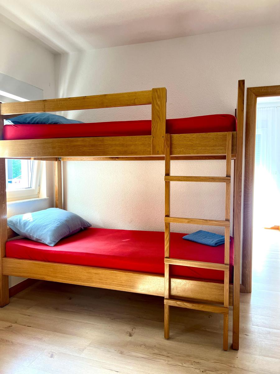 Zimmer mit Holzbett und roten Matratzen. Fenster und Tür sichtbar.