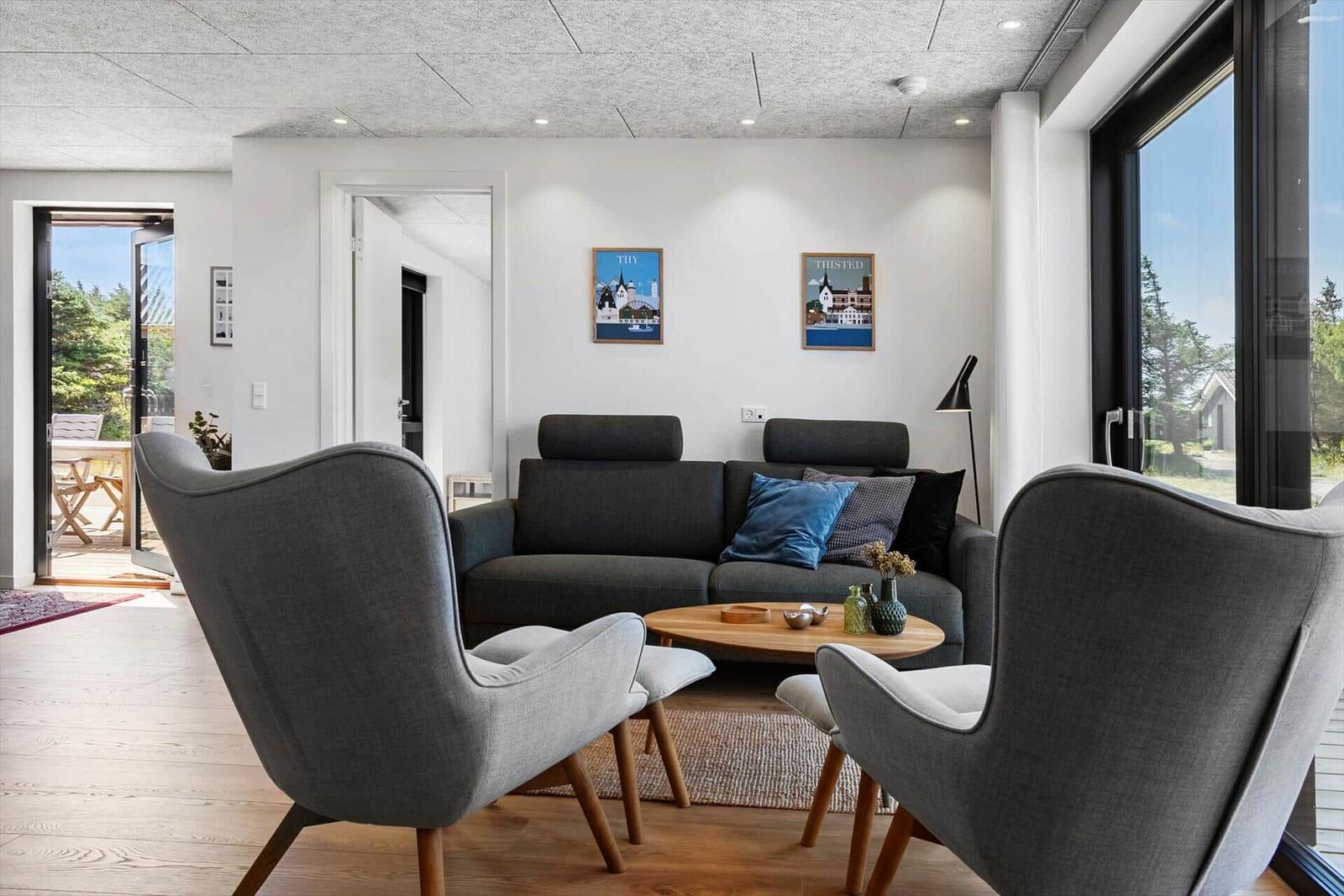 Wohnzimmer mit grauem Sofa, zwei Sesseln und Holztisch. Große Fenster und Türen.