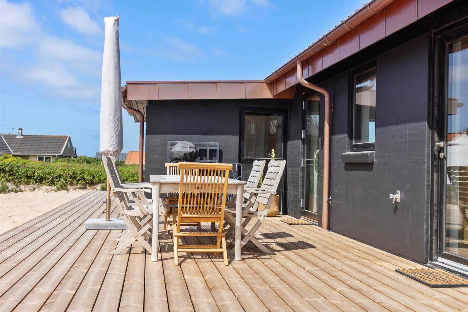 Terrasse mit Holztisch, Stühlen und Sonnenschutz neben einem Haus mit dunkler Fassade.
