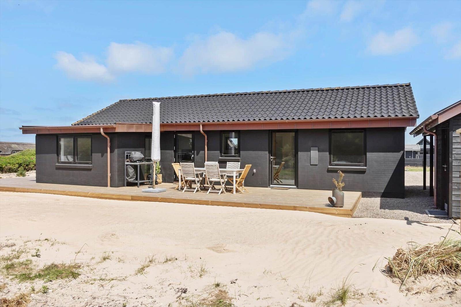 Moderne Ferienhaus mit Terrasse, Grill und Gartenmöbeln an sandigem Grundstück.