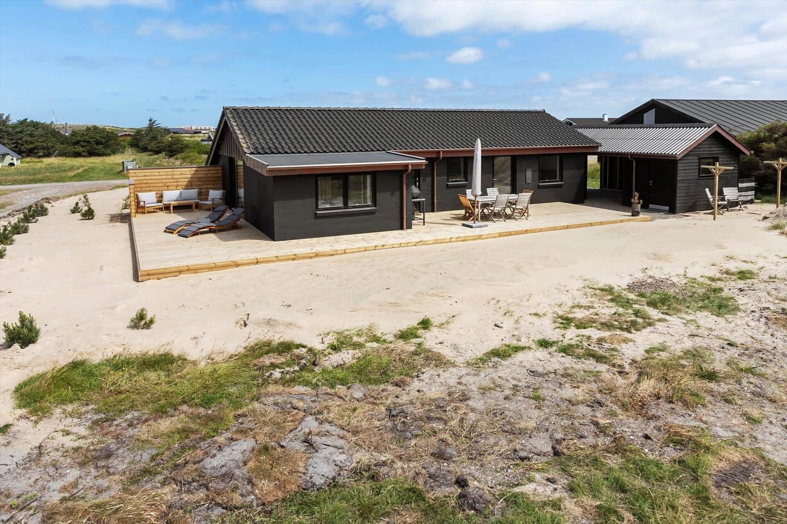 Modernes Ferienhaus mit Terrasse und Gartenmöbeln auf sandigem Grund.