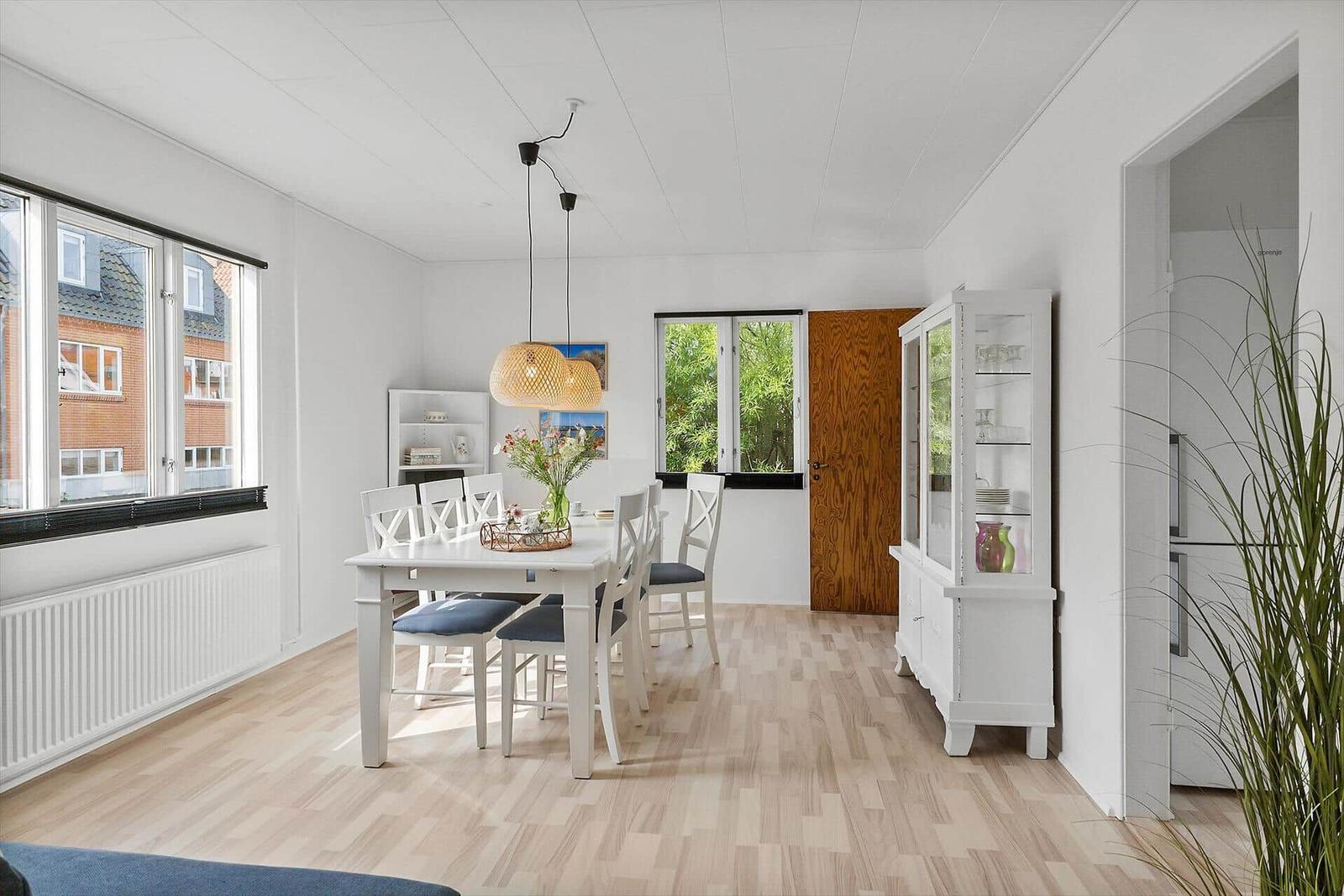 Dining room with white table, chairs, and wooden floor. Windows, cabinet, and plant visible.