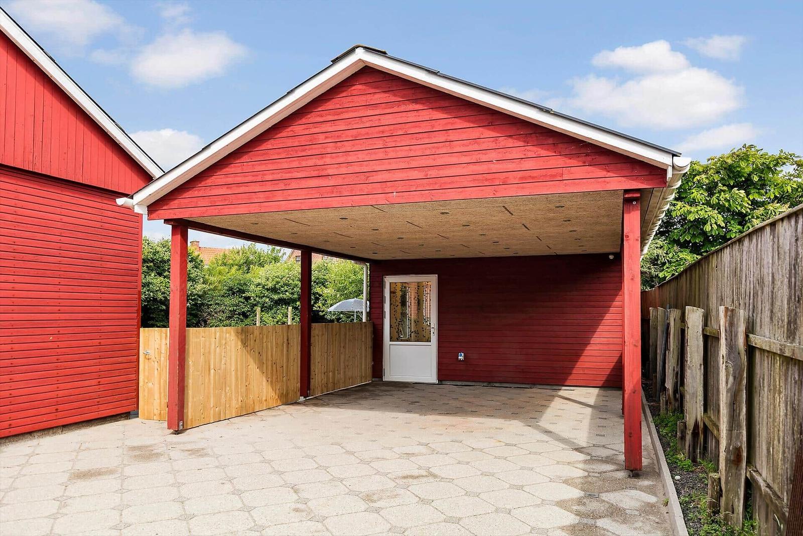 Red carport with wooden cladding and white door. Paved ground and wooden fence.