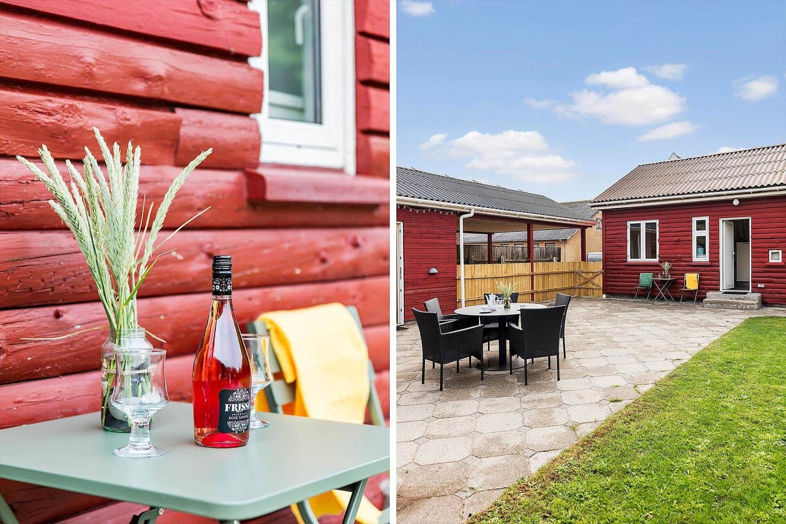 Red wooden house with patio and garden. Table with wine and grass stems.